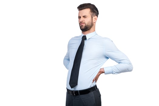 Feeling pain in his back. Frustrated young man in shirt and tie touching his back and keeping eyes closed while standing against white background photo