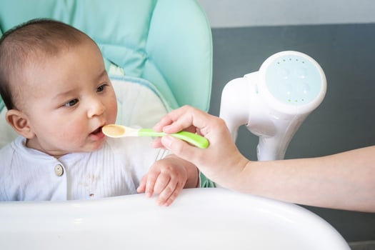 Mom feeds the baby with a spoon of vegetable puree at the children's feeding table. Baby's appetite, healthy nutrition, introduction of complementary foods. Copyspace, mock up photo