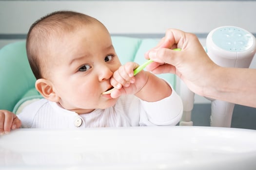 Mom feeds the baby with a spoon of vegetable puree at the children's feeding table. Baby's appetite, healthy nutrition, introduction of complementary foods. Copyspace, mock up photo