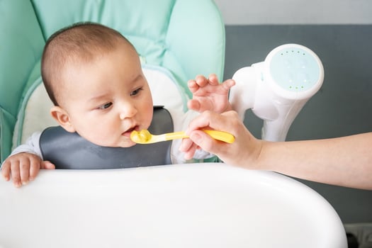 Mom feeds the baby with a spoon of vegetable puree at the children's feeding table. Baby's appetite, healthy nutrition, introduction of complementary foods. Copyspace, mock up photo