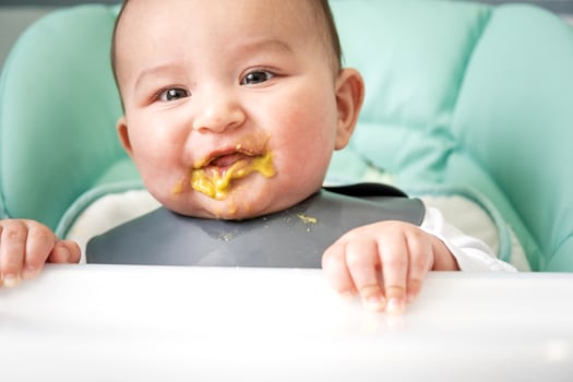 A dirty, joyful baby at the table on a feeding chair got his mouth dirty in vegetable puree. Introduction of complementary foods, the child likes to eat. Portrait close-up photo