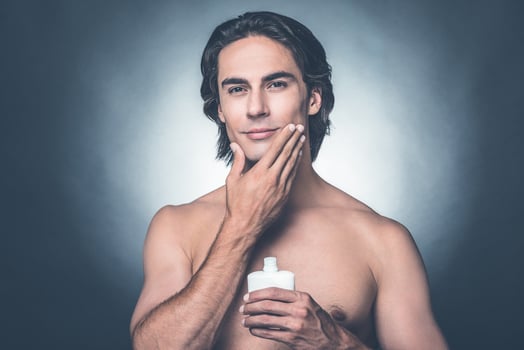 Feeling fresh after shaving. Portrait of handsome young shirtless man looking at camera and applying aftershave lotion on face while standing against grey background photo