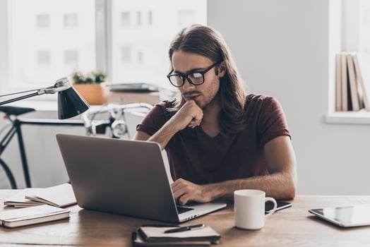 Solutions every day. Thoughtful young man with long hair looking at laptop and holding hand on chin while sitting at his working place in office photo