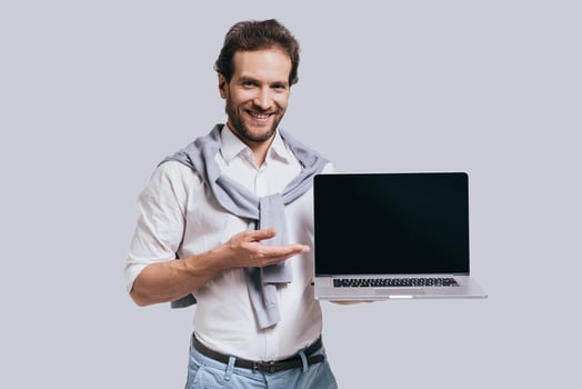 Achieving best results. Beautiful young man in smart casual clothes holding the laptop and smiling while standing against grey background photo