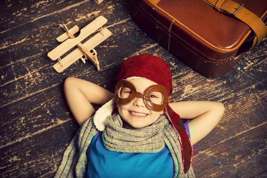 Dreaming of a big sky. Top view of happy little boy in pilot headwear and eyeglasses lying on the hardwood floor and smiling while wooden planer and briefcase laying near him photo