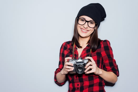 Stylish photographer.  Beautiful young woman in headwear and glasses holding camera and looking at camera while standing against grey background photo