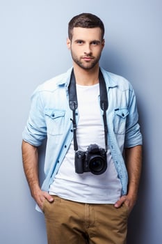 Confident and experienced photographer. Handsome young man with digital camera looking at you while standing against grey background photo