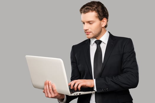 Businessman with laptop. Confident young man in formalwear working on laptop while standing against grey background photo