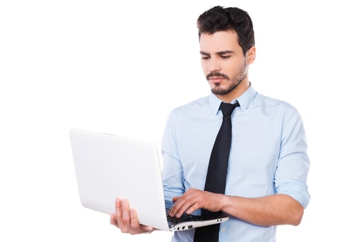 Confident business expert. Handsome young man in shirt and tie working on laptop while standing against white background photo