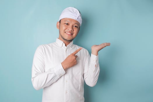 Excited Balinese man wearing udeng or traditional headband and white shirt pointing at the copy space beside him, isolated by blue background photo