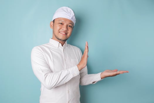 Excited Balinese man wearing udeng or traditional headband and white shirt pointing at the copy space beside him, isolated by blue background photo