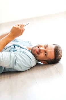 Relaxing at home. Cheerful young man holding mobile phone and looking at camera while lying on the floor at his apartment photo