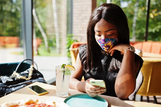 Fashionable african american woman, wear hand made face mask sitting at restaurant, look at mobile phone, while eat pizza. New normal life. photo