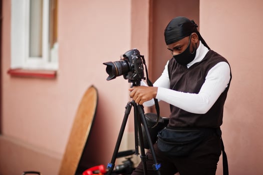 Young professional african american videographer holding professional camera with pro equipment. Afro cameraman wearing black duraq and face protect mask, making a videos. photo