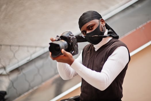 Young professional african american videographer holding professional camera with pro equipment. Afro cameraman wearing black duraq and face protect mask, making a videos. photo