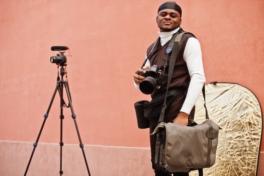 Young professional african american videographer holding professional camera with pro equipment. Afro cameraman wearing black duraq making a videos. photo