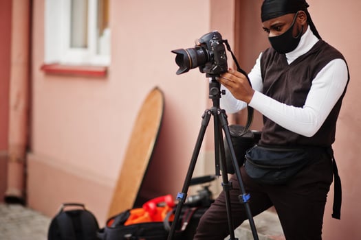 Young professional african american videographer holding professional camera with pro equipment. Afro cameraman wearing black duraq and face protect mask, making a videos. photo