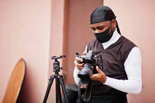Young professional african american videographer holding professional camera with pro equipment. Afro cameraman wearing black duraq and face protect mask, making a videos. photo