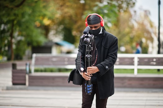 Young professional african american videographer holding professional camera with pro equipment. Afro cameraman wearing black duraq and face protect mask, making a videos. photo