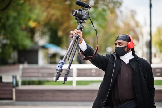 Young professional african american videographer holding professional camera with pro equipment. Afro cameraman wearing black duraq and face protect mask, making a videos. photo
