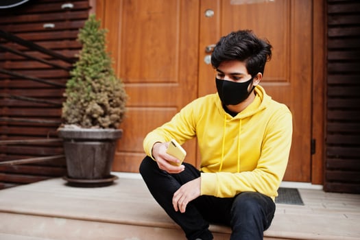 Urban young hipster indian man in a fashionable yellow sweatshirt with mobile phone. Cool south asian guy wear hoodie and black face protect mask, during new normal. photo