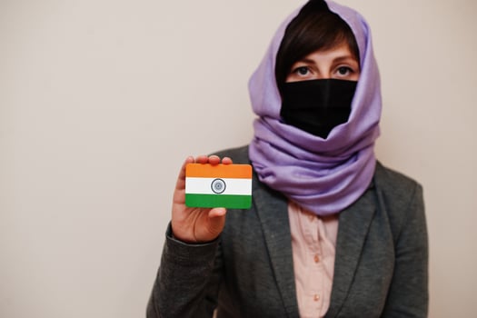 Portrait of young muslim woman wearing formal wear, protect face mask and hijab head scarf, hold India flag card against isolated background. Coronavirus country concept. photo