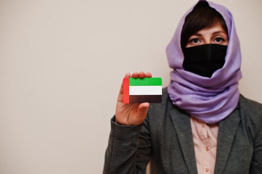 Portrait of young muslim woman wearing formal wear, protect face mask and hijab head scarf, hold United Arab Emirates flag card against isolated background. Coronavirus country concept. photo