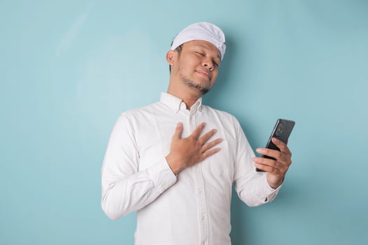 Happy mindful thankful young Balinese man holding phone and hand on chest smiling isolated on blue background feeling no stress, gratitude, mental health balance, peace of mind concept. photo