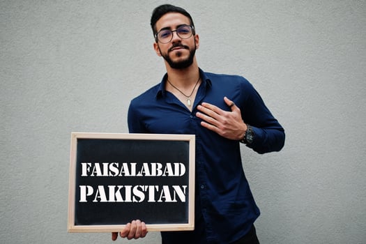 Arab man wear blue shirt and eyeglasses hold board with Faisalabad Pakistan inscription. Largest cities in islamic world concept. photo