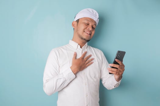 Happy mindful thankful young Balinese man holding phone and hand on chest smiling isolated on blue background feeling no stress, gratitude, mental health balance, peace of mind concept. photo