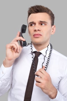 Tired of office work. Depressed young man in shirt and tie talking on the phone and holding telephone wire while standing against grey background photo