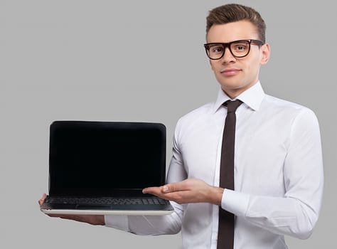 Man with laptop. Handsome young man in shirt and tie holding a laptop and pointing it while standing against grey background photo