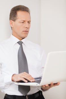 Businessman with laptop. Confident mature man in shirt and tie working on laptop while standing near the wall photo