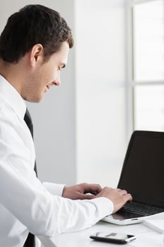 Working with pleasure. Side view of smiling young man in formalwear working on laptop while sitting at his working place photo