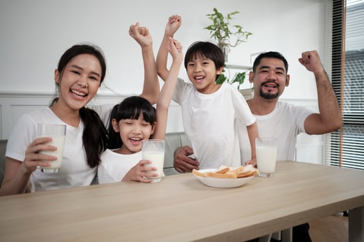 A Healthy Asian Thai family, little children, and young parents drink fresh white milk in glass and bread joy together at a dining table in morning, wellness nutrition home breakfast meal lifestyle. photo