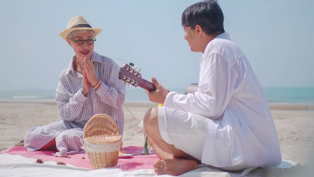 Asian retired couple relaxing by the sea in summer. A senior man playing guitar while having a picnic with his partner on the beach. photo