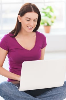 Surfing the net at home. Attractive young woman working on laptop and smiling while sitting on the floor at her apartment photo