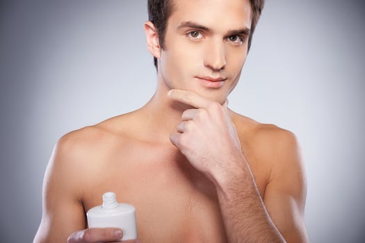 No irritation. Handsome young shirtless man applying cream at his face and looking at camera while standing against grey background photo