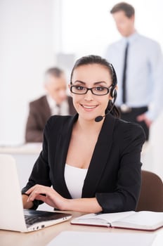Customer service representative at work. Attractive young woman in formalwear adjusting her headset and smiling while people working on background photo