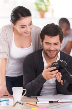 Good shots Two co-workers discussing something while man holding camera and smiling with people working on background photo
