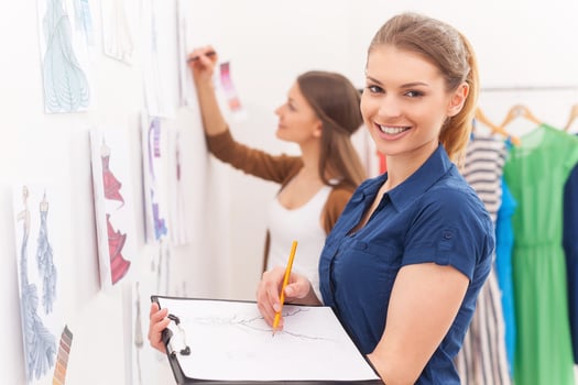 Fashion designers sketching. Beautiful female fashion designer holding clipboard with sketches and smiling while her colleague standing on background photo