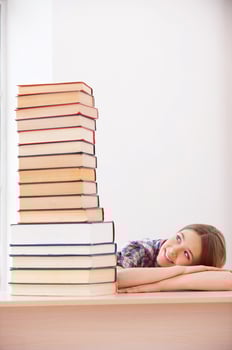 She loves studying. Cheerful teenage girl looking at the top of the big stack of books laying on the desk photo
