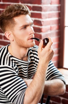 Man smoking pipe. Handsome young man in striped shirt smoking pipe and looking away while sitting on the chair photo