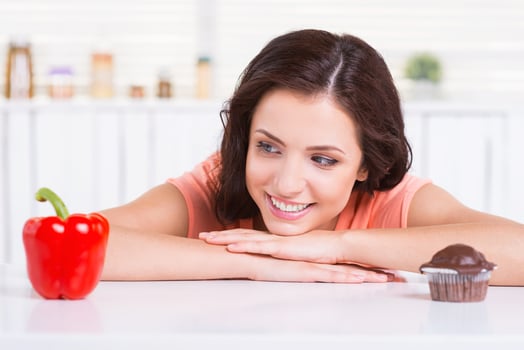Making her choice. Cheerful young woman choosing what to eat while leaning at the kitchen table with chocolate muffin and fresh pepper laying on it photo