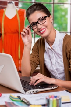 Beautiful fashion expert. Beautiful young fashion designer adjusting her glasses and smiling at camera while sitting at her working place photo