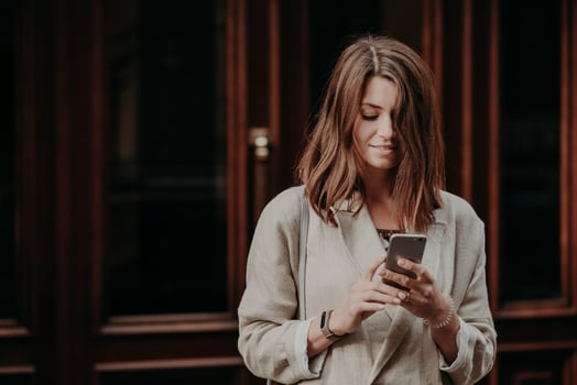 Pleased delighted female youngster watches video on smart phone, reads banking notification, dressed in white raincoat, poses against building interior. People, lifestyle and chatting concept photo