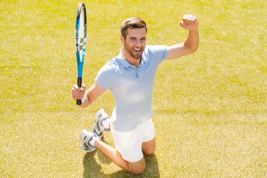 I won Top view of happy young man in polo shirt holding tennis racket and gesturing while kneeling on tennis court photo