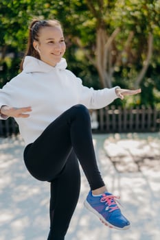 Fitness sport healthy lifestyle concept. Pleased sporty woman tries to balance on one leg keeps knee raised has interval training or cardio workout wears white sweatshirt leggings and sneakers photo