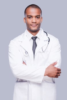 Confident male doctor. Confident African doctor looking at camera and keeping arms crossed while standing against grey background photo