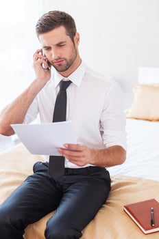 No time for rest. Confident young man in shier and tie talking on the mobile phone and holding document while sitting on the bed in hotel room photo